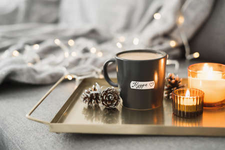 Hygge concept: black mug with hot coffee served on a metal tray, burning candles, pine cones as a decor and christmas lights on background