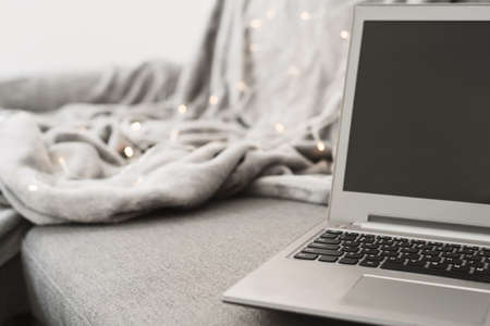 Laptop computer standing at gray sofa, blanket and decorative lights as background. Work from home conceptの写真素材