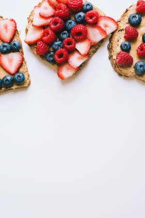 Vertical background with peanut butter toasts with seasonal berries. Brown bread with creamy spread, strawberry, blueberry and raspberry on white table. Healthy snackの写真素材