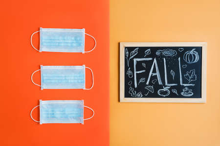 Medical Face masks and blackboard with handwritten text "FALL" on bright orange and yellow background. Autumn season during Covid 19 pandemicの写真素材