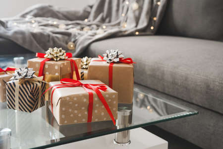 Gift boxes wrapped in craft paper on glass coffee table in the living room, sofa bed with blanket and decorative lights as background. Christmas presentsの写真素材