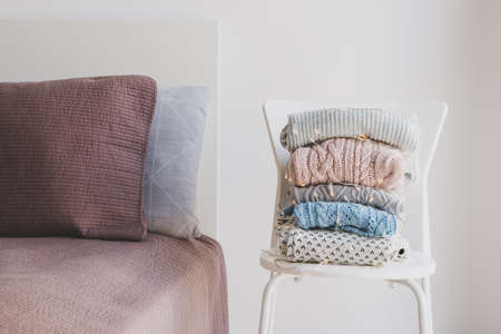 Pile of warm woolen sweaters wrapped in decorative lights on white chair and clean and neat bed. Knitwear care and storage conceptの写真素材