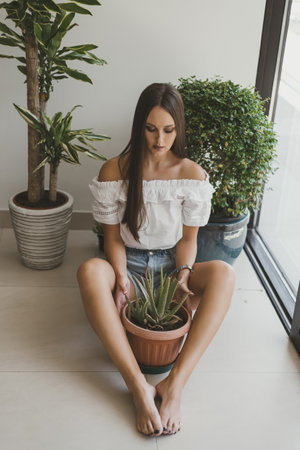 Young woman sitting on the floor in her apartment and holding a pot with Aloe Vera plant. Indoor garden care. Houseplantsの写真素材
