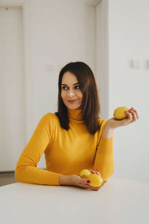 Young European woman wearing a bright yellow turtleneck sweater sitting at the dining table and holding lemonsの写真素材
