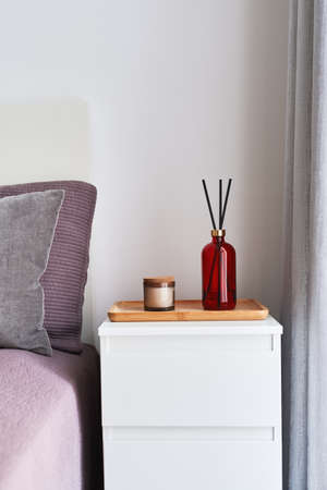 Bottle of fragrance diffuser with sticks and aroma candle on a bedside table in a bedroom. Interior...
