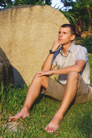 Relaxed attractive smiling man sit in a shirt, shorts and hours near a large stone.の写真素材