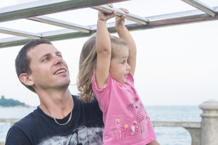Dad and daughter playing promenade by the sea. young man, little girl hanging on the barの写真素材