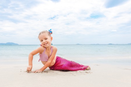 Little beautiful girl dressed in swimsuit as a mermaid sits on the seashoreの写真素材