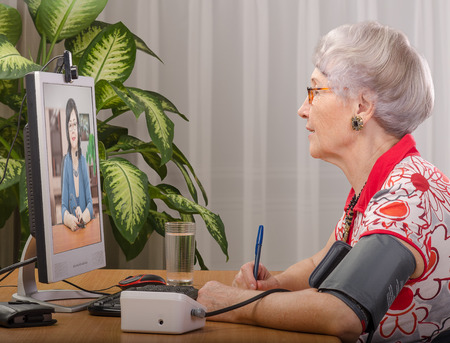 Old grey haired woman is measuring blood pressure while virtual doctor consults her on monitor screenの写真素材