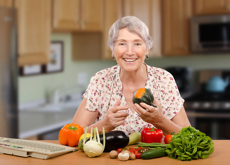 Smiling senior woman is sitting at the table with fresh vegetables. She is online client of dietitian. Nutritionist helps her with menu planningの写真素材
