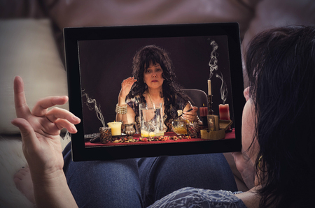 Woman on a sofa with tablet computer has online conversation with psychic doing egg divination. Black haired fortune teller is reading a shapes of egg whites in  touchscreenの写真素材