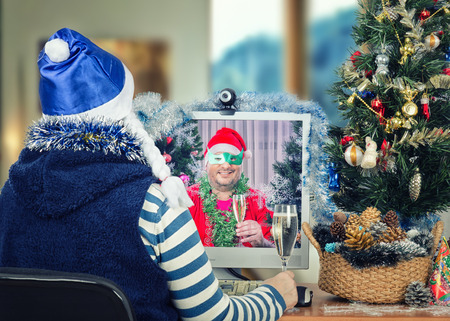 Woman in blue hat with white pigtails sits at the desk looks at monitor and drinks sparkling white wine online with Santa Claus wearing green domino eye maskの写真素材