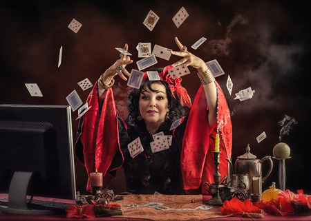 Mature Gypsy fortune-teller sitting opposite monitor tossing playing cards up to predict future for her online customerの写真素材