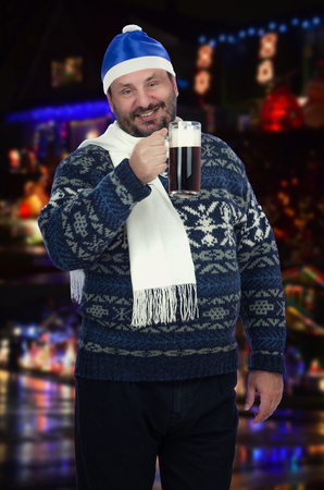 Bearded man in Santa Claus blue hat is dark beer lover. He is going to chug a pitcher as fast as he can. Middle-aged man wearing winter sweater, white scarf and posing on street sparkling blurred backgroundの写真素材