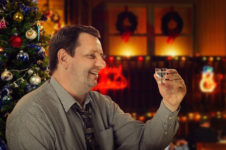 Smiling mature man makes a toast for Christmas Eve. Side portrait of friendly man holding vodka shot. He sits in a cafe on holiday blurred  backgroundの写真素材