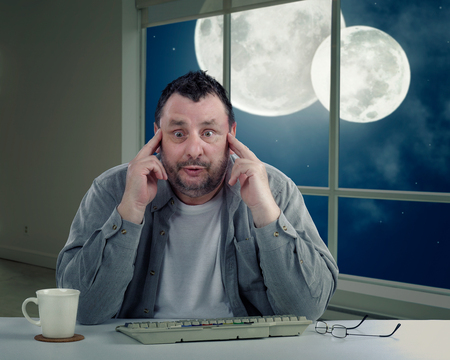 Man with bulging eyeballs is frightened to turn around to look again at  two moons on night sky in the window. Scared middle-aged man holds his head with fingers. Man has never seen double moonの写真素材