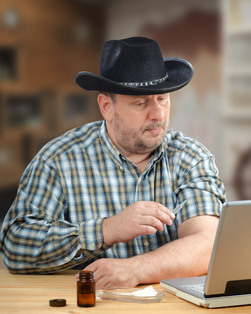 Owner of farm in remote area learning how to get vaccinated against the flu for himself during virtual telemedicine consultation. Cowboy in black hat sits with syringe in front of laptopの写真素材