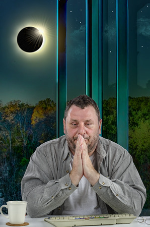 Mature man is very worried because of the solar eclipse. Solar eclipse affected his mood. He puts his palms in praying gesture and looks down sitting at office desk with keyboardの写真素材