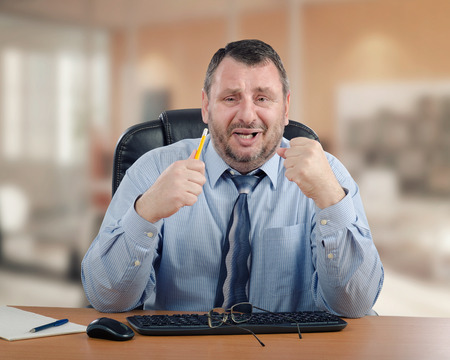 Panicked manager clenches his hands because he cannot make the right decision. Mature man wearing blue shirt and striped tie is ready to cry. He sits at wooden office deskの写真素材