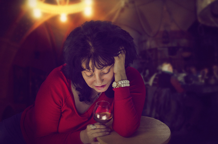 Alcoholic mature woman in red jumper drinks alone leaning on wooden stool in cafe. Black-haired woman is looking down into a glass of brandy sadly. Dark indoors blurred backgroundの写真素材