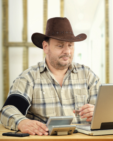 Cowboy makes self-monitoring of blood pressure with telehealth support. Middle aged farmer wearing plaid shirt and brown hat sits in front of computer. Man suffers hypertension measures BP levels at regular intervals and transmit it to telemedicine headquarterの写真素材