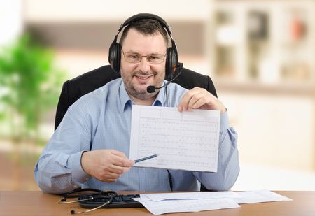 Positive virtual male doctor goes over printed heart-rate graph looking at the camera. Middle-aged man in headset wearing blue shirt sits at wooden office desk. Indoors blurred backgroundの写真素材