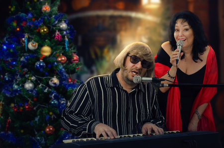 Man and woman sing duets at Christmas songs. Man in light yellow wig plays electric piano. Mature woman in big red scarf is singing nearby. Holiday indoors blurred backgroundの写真素材