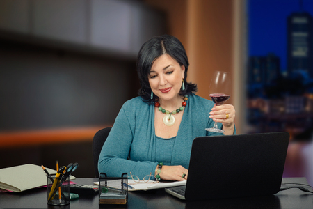 Cute middle aged business woman in official merino light blue suit relaxes chatting online after the hard day. She holds glass of red wine while listening to a chatter partner.の写真素材