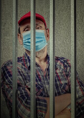 Mature man is placed in a pre-trial detention cell for COVID-19 quarantine violation. He is in a face mask looking down through the prison bars.の写真素材