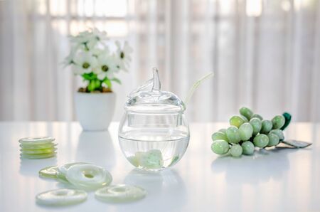 Polished small jade stones are located inside of a glass jar with crystalline water. It is making crystal charged water at home. There are donuts carved grapes and white flowers on the table.の写真素材