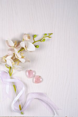Plastic orchid sprig with the greenish stem wrapped by silk ribbons and two rose quartz hearts lay on a light gray wooden background. Top view, copy space.の写真素材
