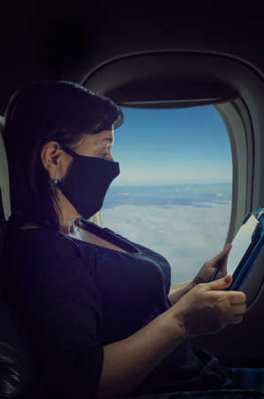 A mature woman in a protective mask is flying by plane during the pandemic. She is reading something on the tablet. Night shot on the window background.の写真素材