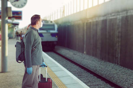 Man in a medical mask and blue rubber gloves is waiting for a train. He stands on the platform with baggage.の写真素材