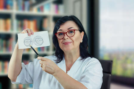 Middle-aged friendly eye doctor in white uniform looking at the camera. She shows a diagram of the normal field of view (FOV).  Woman has black hair and wears glasses.の写真素材