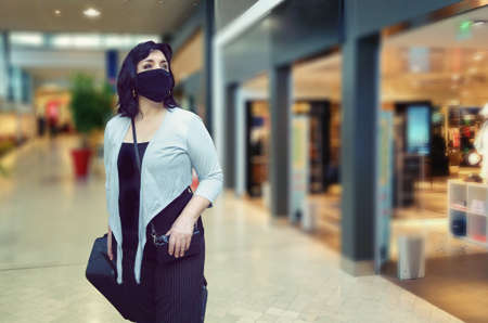 Adult woman in a black mask glances at the storefronts while walking through an airport. She rolls a big black suitcase on wheels.の写真素材