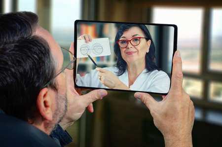 Mature man sits at home holding a tablet in his hands. He carefully looks at the telemedicine doctor on the touch screen. GP recommends his taking field of view (FOV) test as soon as possible.の写真素材