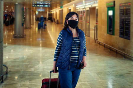 Woman in a black protective face mask goes through the arrival or departure hall of an airport. She is dressed in dark blue clothes and rolls a suitcase on wheels.の写真素材