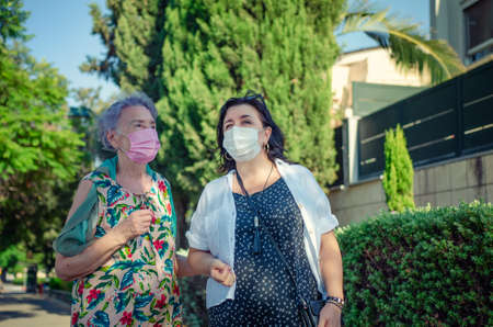 Caregiver or nurse and a senior adult woman stroll in a green street during coronavirus. Both are wearing face protective masks.の写真素材