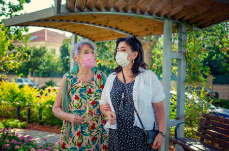 Companion or volunteer and a senior adult woman wearing a protective mask during coronavirus. They are walking in the park next to a house.の写真素材