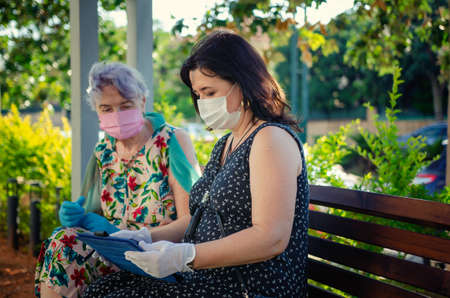 A kind volunteer teaches a senior adult woman how to enjoy a digital tablet. Both in protective face masks sitting on a park bench. Summer sunny day.の写真素材