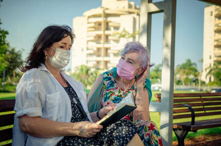 An elderly woman listens very carefully to a kind volunteer reading book aloud. It is providing emotional support for old people.  Both in protective face masks sitting on a park bench.の写真素材