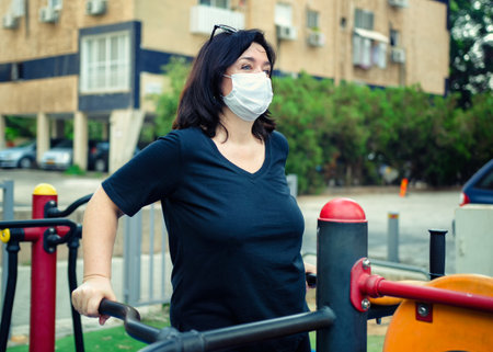 Mature woman in a face mask likes exercising at an outdoor fitness station near her residence. She...