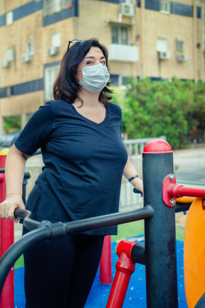Despite the coronavirus, the mature woman continues to play sports in the outdoor fitness field near her home. She puts on a face mask for protection.の写真素材