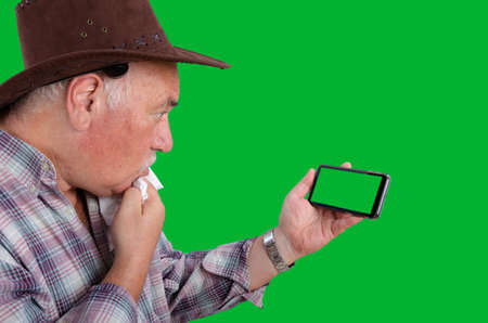 Old rancher in a brown hat checkered shirt staring in confusion at a chroma key screen on a smartphone. Close up side shot on a green background.の写真素材