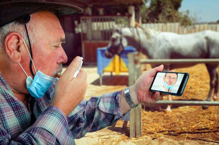 An old farmer, choking with coughs, speaks to a telemedicine doctor on his smartphone.の写真素材