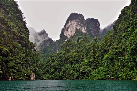 Unique Cheo Lan Lake on a rainy day. On the emerald water of the lake, ripples from raindrops. On the shore there are unusual high cliffs covered with lush tropical vegetation. The fog hides the peaks of the mountains. Mysteriously.の写真素材