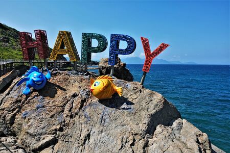 Installation Happy. On the rock are large multi-colored volumetric letters filled with empty metal cans of drinks. Nearby are bright plastic fish. Background - ocean, blue sky and mountains silhouettes.の写真素材