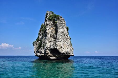 Lonely island in the Andaman Sea. A rock in the form of a pyramid rises high above the turquoise water. There are few plants on top. Reflection in water. The background is a clear bright blue sky.の写真素材