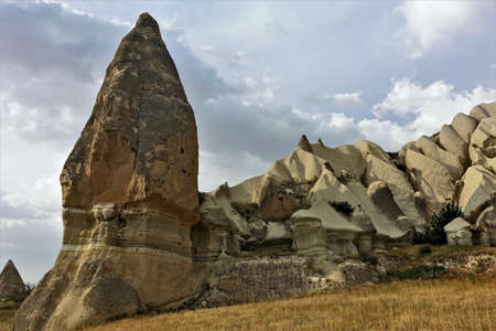 Strange rocks of Cappadocia. Against the background of the cloudy sky, unusual pointed mountains of a fanciful form rise. Color - different shades of sand. Yellow dry grass grows around. UNESCOのeditorial素材