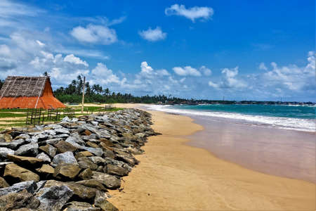 Summer, beach, ocean. Aquamarine waves leave a white foam on the yellow sand. Stones lie along the shore, and there is a red wooden hut. Silhouettes of palm trees against the blue cloudy sky.Sri Lankaの写真素材